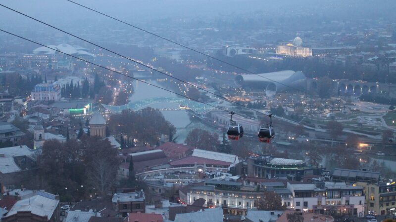 Panoramic twilight view of Tbilisi, Georgia, showing the illuminated Peace Bridge over the Kura River, cable cars in motion, and the cityscape fading into mist. Key landmarks like the Rike Park glass structures and the Presidential Palace are visible in the distance.