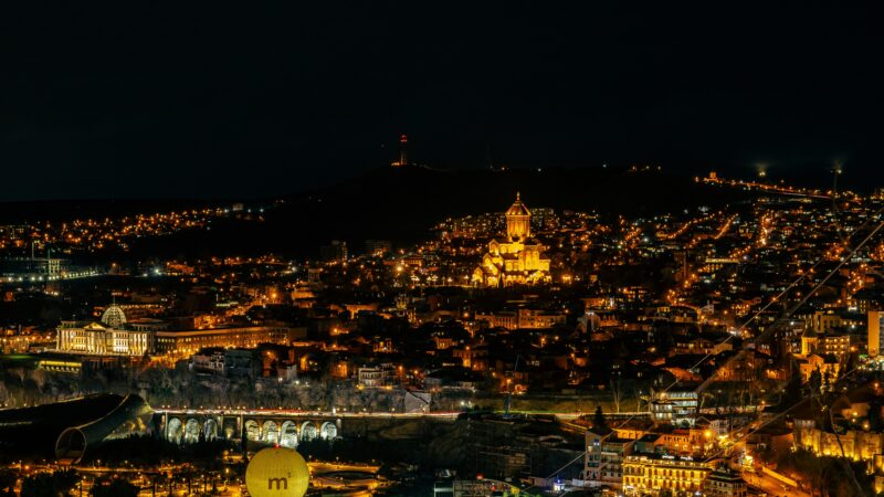 A panoramic nighttime view of Tbilisi, Georgia, showcasing its illuminated skyline. Key landmarks include the brightly lit Sameba Cathedral (Holy Trinity Cathedral) on a hill, the distinctive yellow "M²" sphere of the Museum of Modern Art in the foreground, and the arched Metekhi Bridge with streaks of traffic light. Thin lines of the city's cable car system crisscross the scene, connecting the densely packed, warmly lit buildings that cascade down the hillsides against a dark sky.