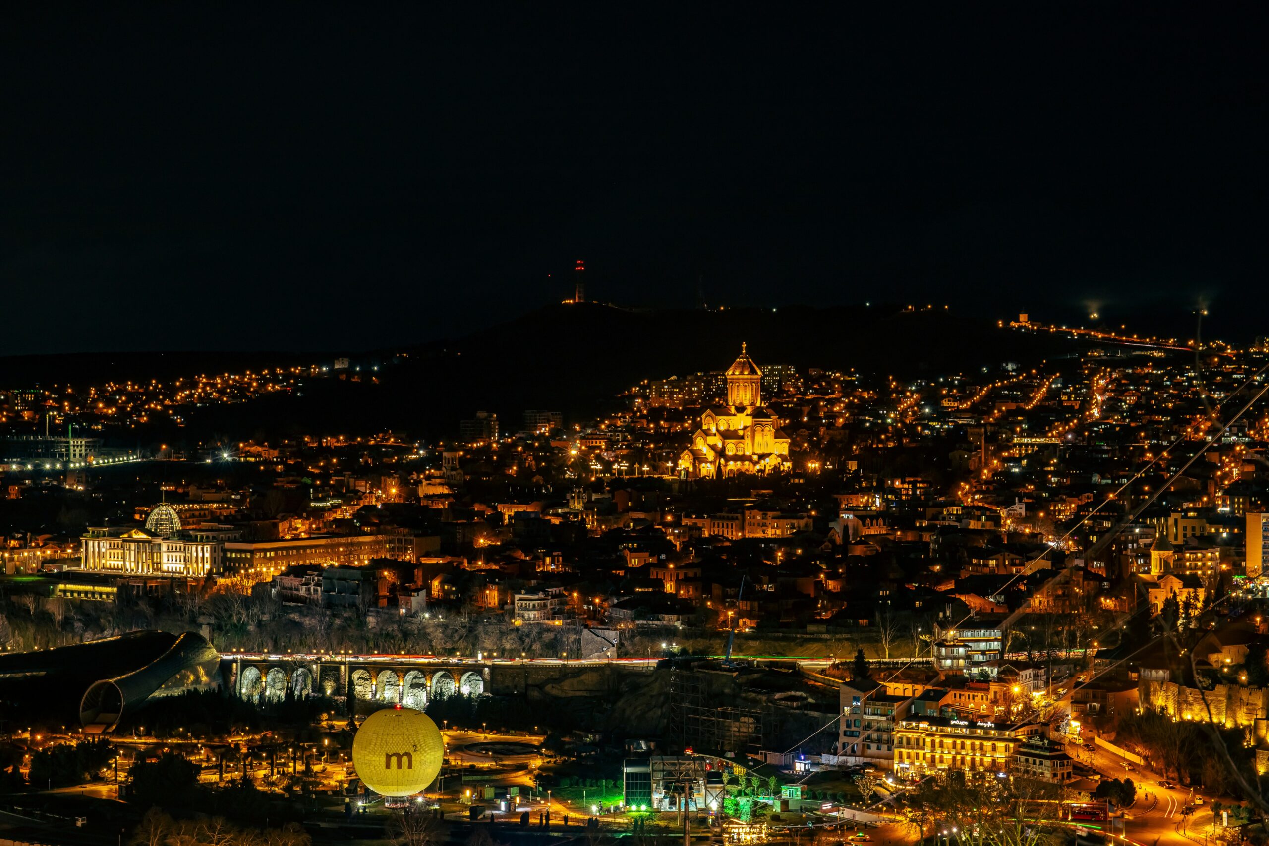 A panoramic nighttime view of Tbilisi, Georgia, showcasing its illuminated skyline. Key landmarks include the brightly lit Sameba Cathedral (Holy Trinity Cathedral) on a hill, the distinctive yellow "M²" sphere of the Museum of Modern Art in the foreground, and the arched Metekhi Bridge with streaks of traffic light. Thin lines of the city's cable car system crisscross the scene, connecting the densely packed, warmly lit buildings that cascade down the hillsides against a dark sky.