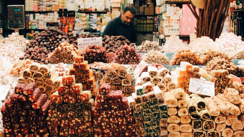 A vendor at a bustling Istanbul market stall displays an abundant variety of traditional Turkish sweets and dried fruits, including dates and honey-flavored Turkish Delight (Ballı Lokumlar).