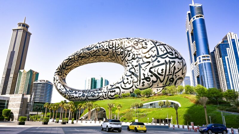 An exterior view of the Museum of the Future in Dubai, featuring its unique torus shape adorned with Arabic calligraphy, surrounded by green landscaping and towering skyscrapers like the Emirates Towers.