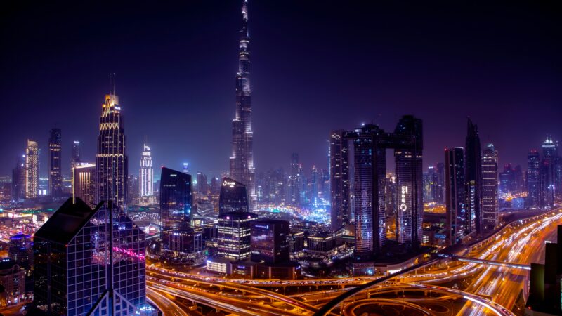 An elevated nighttime panorama of Downtown Dubai, showcasing the towering, illuminated Burj Khalifa against a dark sky, with the bustling Sheikh Zayed Road highway creating streaks of light below.