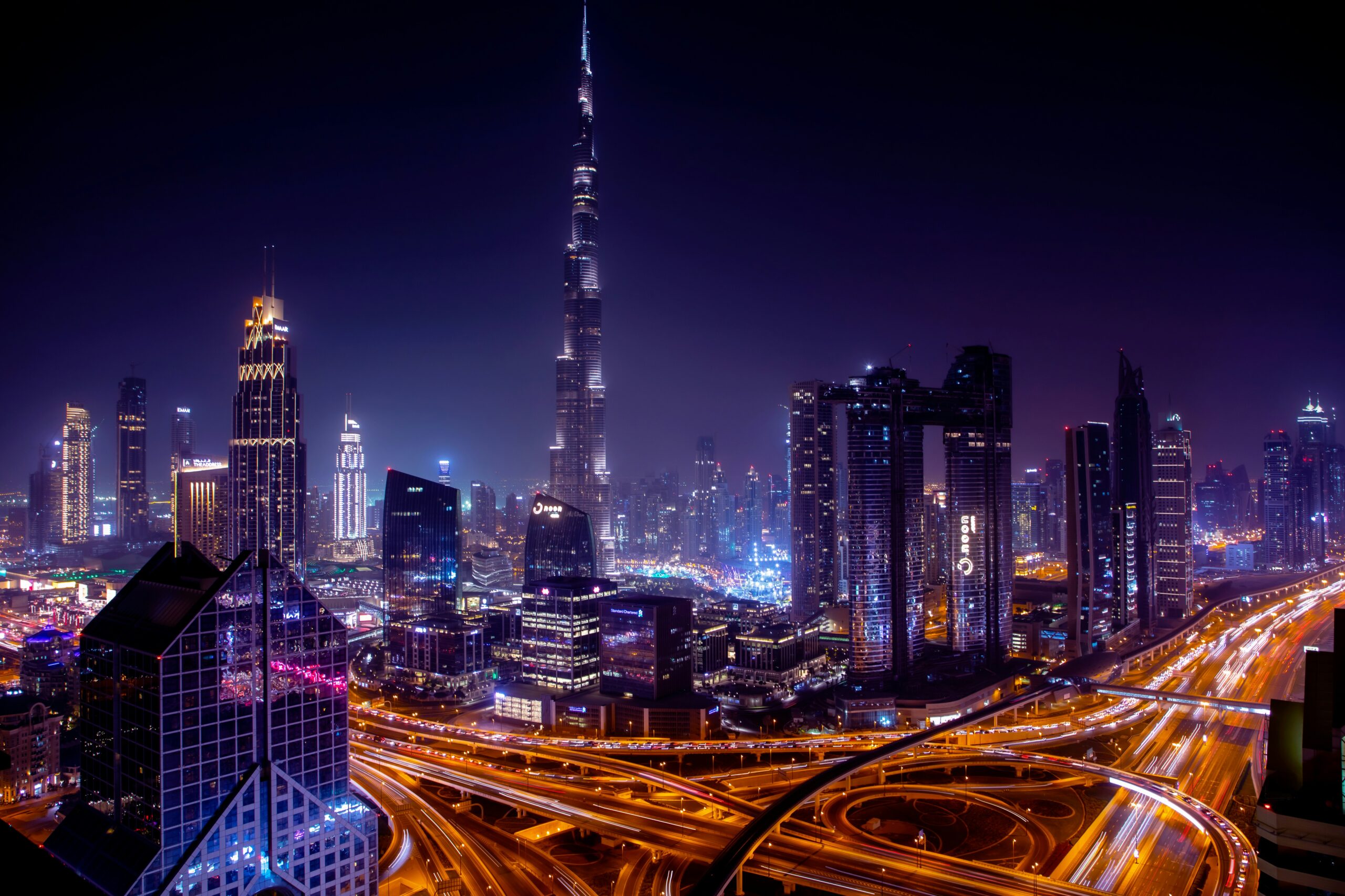 An elevated nighttime panorama of Downtown Dubai, showcasing the towering, illuminated Burj Khalifa against a dark sky, with the bustling Sheikh Zayed Road highway creating streaks of light below.