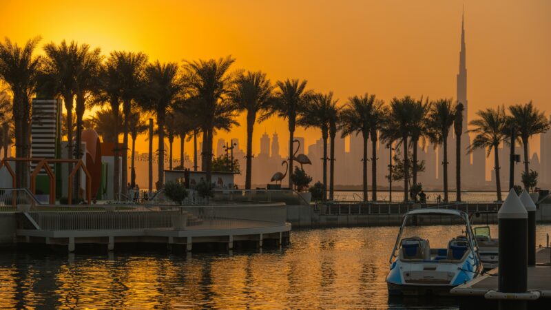 Warm, golden sunset over Dubai Marina. Silhouetted palm trees frame the view of the city skyline, including the Burj Khalifa, with calm water reflecting the light and boats moored nearby.